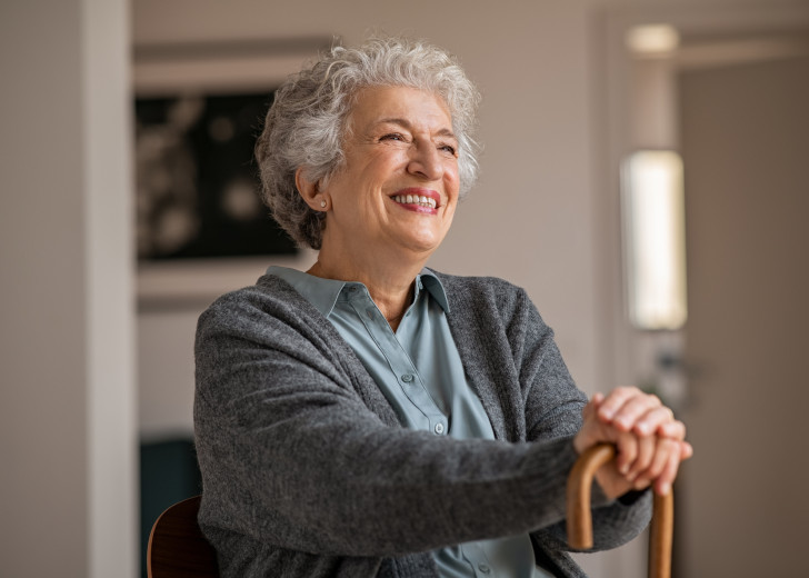 Retired senior woman laughing with her wooden walking stick while relaxing at home. Happy smiling old woman holding walking cane and looking through the window with positivity.