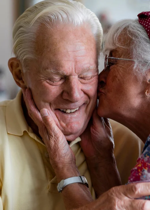 Senior couple at home in London. The wife is kissing her husband's cheek while he laughs with his eyes closed.