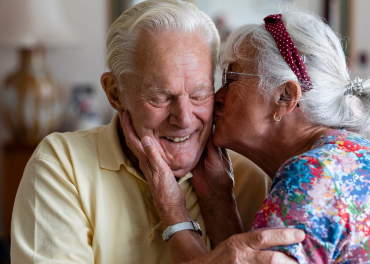 Senior couple at home in London. The wife is kissing her husband's cheek while he laughs with his eyes closed.