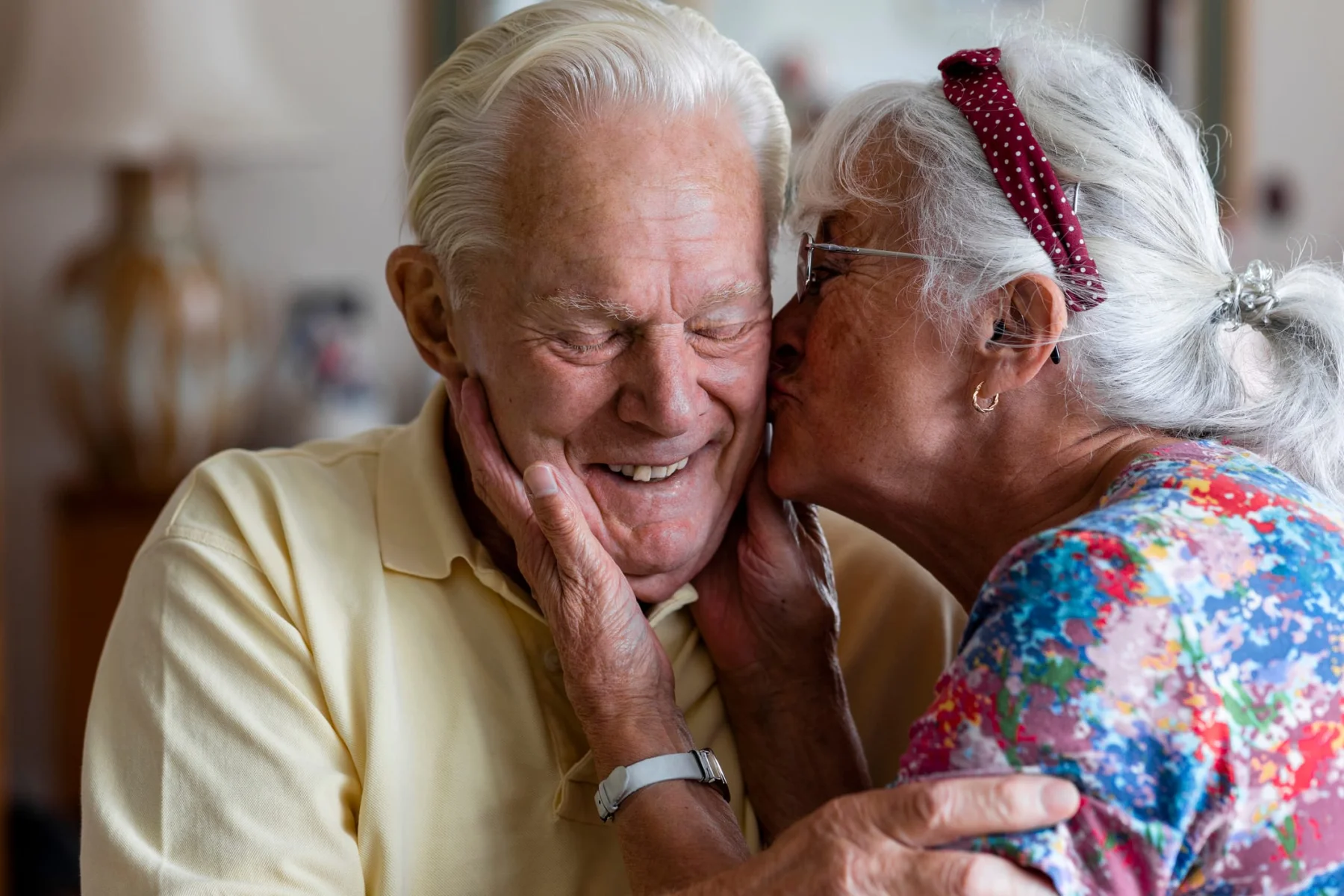 Senior couple at home in London. The wife is kissing her husband's cheek while he laughs with his eyes closed.